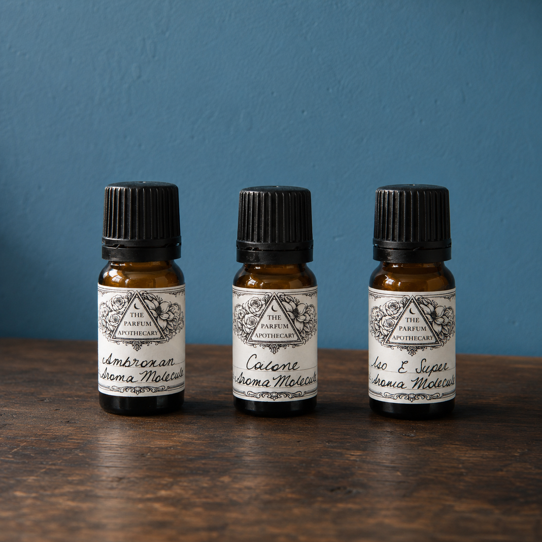 Three modern perfume ingredients in small amber bottles with black caps on a wooden surface against a blue wall.